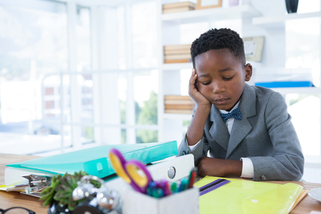 Tired kid businessman with hand on chin sitting at desk in officeの写真素材