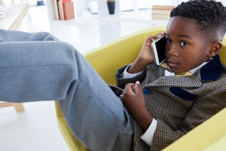 Close up of kid businessman talking on smartphone while sitting on armchair in officeの写真素材