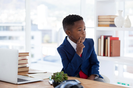 Thoughtful kid businessman with hand on chin looking away sitting at desk if officeの写真素材