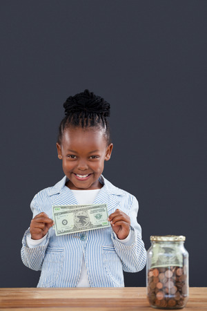 Portrait of smiling businesswoman with jar at desk showing paper currency against gray wall in officeの写真素材