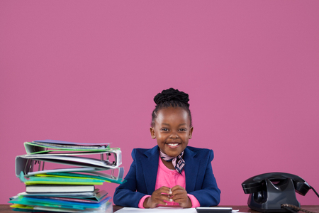 Portrait of smiling businesswoman with files and telephone sitting at desk against pink wall in officeの写真素材