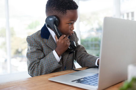 Businessman with laptop talking on telephone while sitting at desk in officeの写真素材