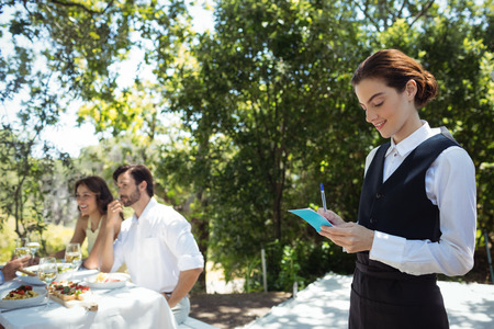 Smiling waiter writing order on notepad while friends dining in background