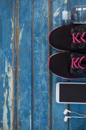 Overhead view of sports shoes by mobile phone with in-ear headphones and water bottle on blue wooden tableの写真素材