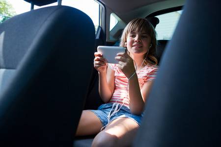 Happy teenage girl in headphones using mobile phone in the back seat of carの写真素材