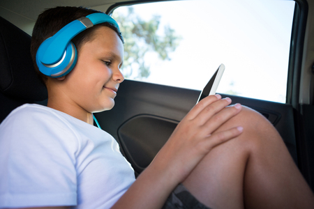 Happy teenage boy with headphones using digital tablet in the back seat of carの写真素材