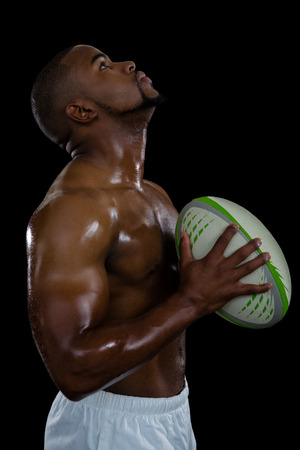 Side view of shirtless male athlete looking up holding rugby ball against black backgroundの写真素材