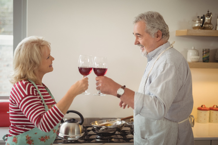 Senior couple toasting glasses of wine while preparing food in kitchenの写真素材
