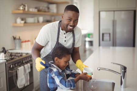Portrait of smiling father and son cleaning cup in kitchen at homeの写真素材