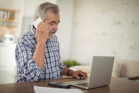 Worried senior man taking on phone while using laptop at homeの写真素材