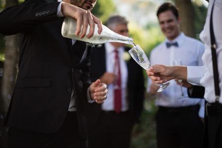 Mid section of groom pouring champagne into his friends glasses in parkの写真素材