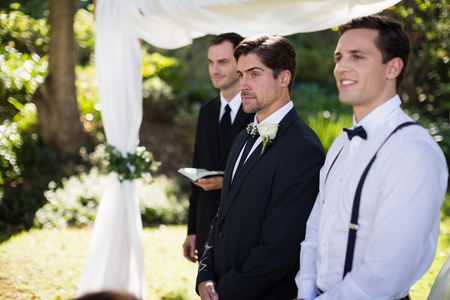 Thoughtful groom standing with waiter and groomsman in parkの写真素材
