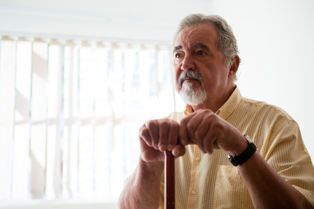 Thougtful senior man looking away while holding walking cane in nursing homeの写真素材