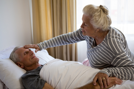 Senior woman sitting by husband sleeping on bed in nursing homeの写真素材
