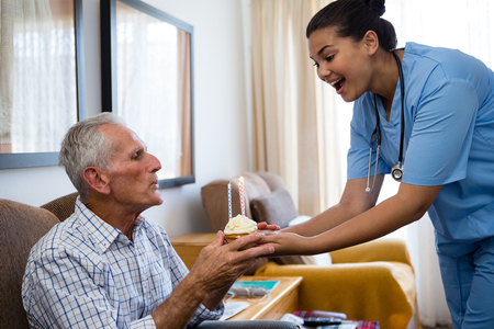 Senior man blowing candles of cup cake being held by female doctor in nursing homeの写真素材