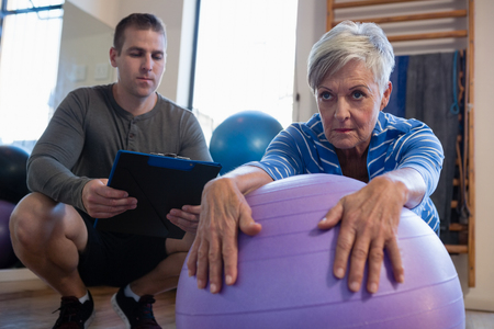 Physiotherapist maintaining record of senior woman performing exercise on fitness ball at clinicの写真素材