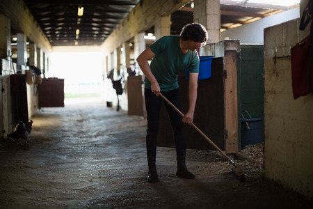 Young man using broom to clean the stableの写真素材