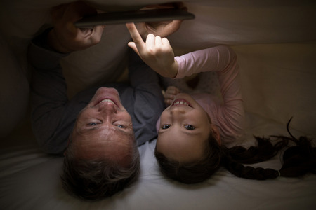 Portrait of smiling father and daughter with digital tablet under blanket on bed in bedroomの写真素材