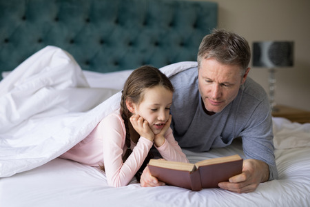 Father and daughter reading book on bed in bedroomの写真素材