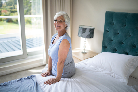 Portrait of smiling senior woman sitting on bed in bedroomの写真素材