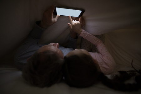 Father and daughter using digital tablet under blanket on bed in bedroomの写真素材