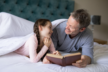 Smiling father and daughter reading book on bed in bedroomの写真素材