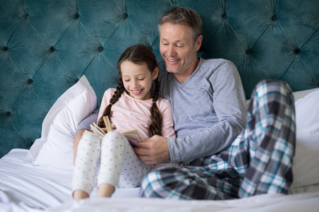 Smiling father and daughter reading book on bed in bedroomの写真素材