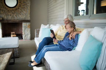 Smiling granddaughter and grandmother using digital tablet on sofa in living roomの写真素材