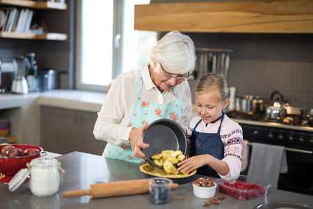 Grandmother and granddaughter adding fresh cut apples to the crust while making apple pie in the kitchenの写真素材