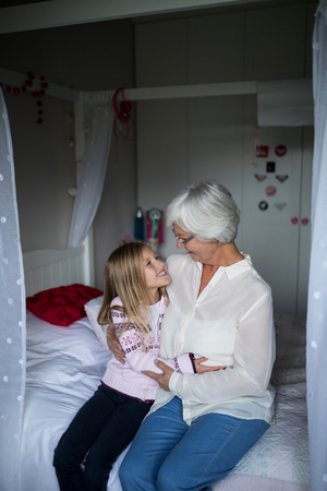 Smiling grandmother and granddaughter embracing each other on bed in bedroomの写真素材