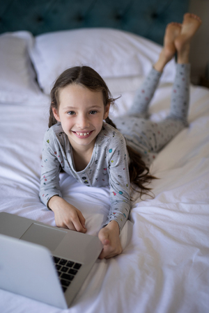 Portrait of smiling girl lying on bed with laptop in bedroomの写真素材
