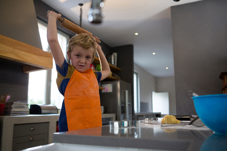 Portrait of boy holding rolling pin with arms raised in kitchen at homeの写真素材