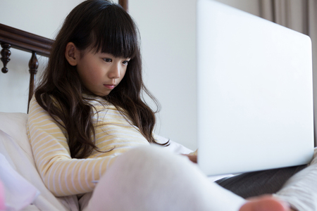 Girl with long hair using laptop while sitting on bed at homeの写真素材