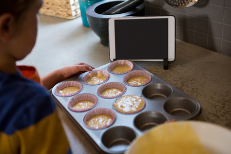 Boy holding muffin tin with cupcake holders by digital tablet at kitchen counterの写真素材