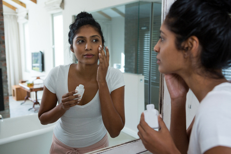 Young woman applying lotion reflecting on mirror in bathroomの写真素材