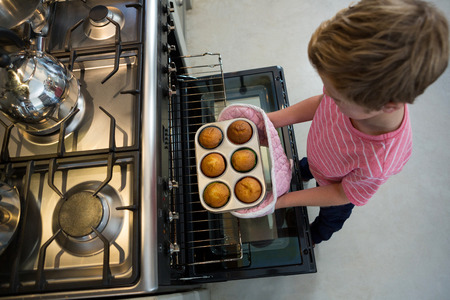 High angle view of boy holding muffin tin by oven in kitchen