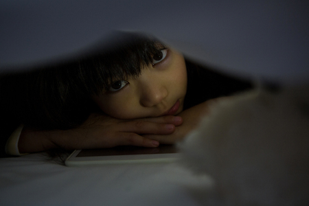 Portrait of girl with digital tablet relaxing below blanket on bedの写真素材
