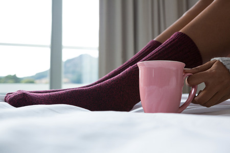 Low section of woman with coffee cup relaxing on bed at homeの写真素材