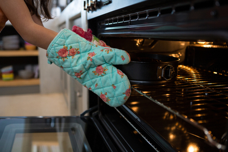 Cropped hands of girl wearing glove putting container with cake in oven at kitchenの写真素材