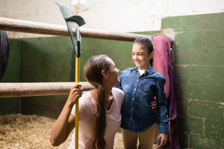 Smiling female jockey holding rake while talking to sister in stableの写真素材