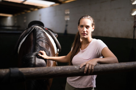 Portrait of young female jockey standing by horse in stableの写真素材