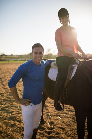 Portrait of trainer standing by young woman sitting on horse at barnの写真素材