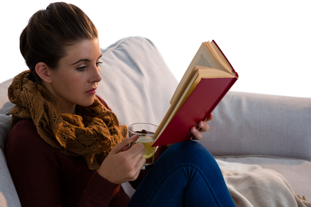 Young woman reading book while having tea on sofa against white backgroundの写真素材