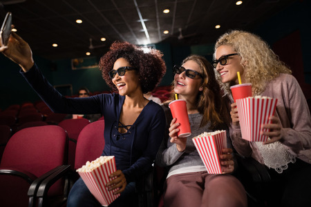 Female friends taking selfie with mobile phone while watching movie in theatreの写真素材