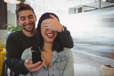 Young man covering girlfriend eyes while gifting ring at cafeの写真素材