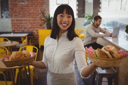 Portrait of waitress holding baskets with sandwiches while businessman using laptop at counter in cafeの写真素材