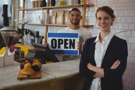 Portrait of confident female owner with waiter holding open sign at counter in cafeの写真素材