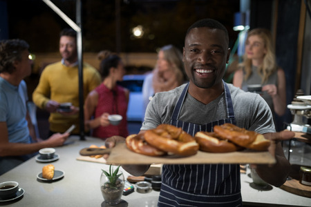 Portrait of happy waiter holding sweet food in wooden tray at cafe counterの写真素材