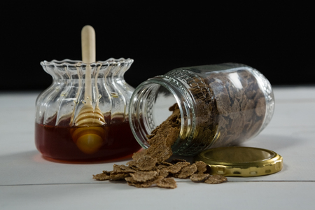 Jar of honey and wheat flakes spilling out of bottle on white background