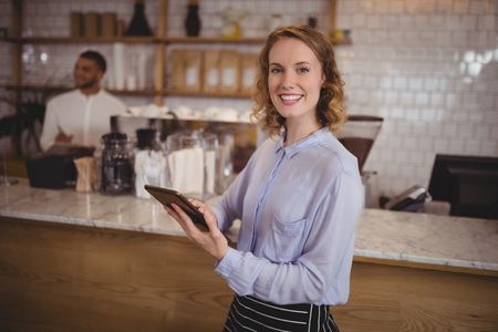 Smiling young waitress using digital tablet while standing by counter at coffee shopの写真素材